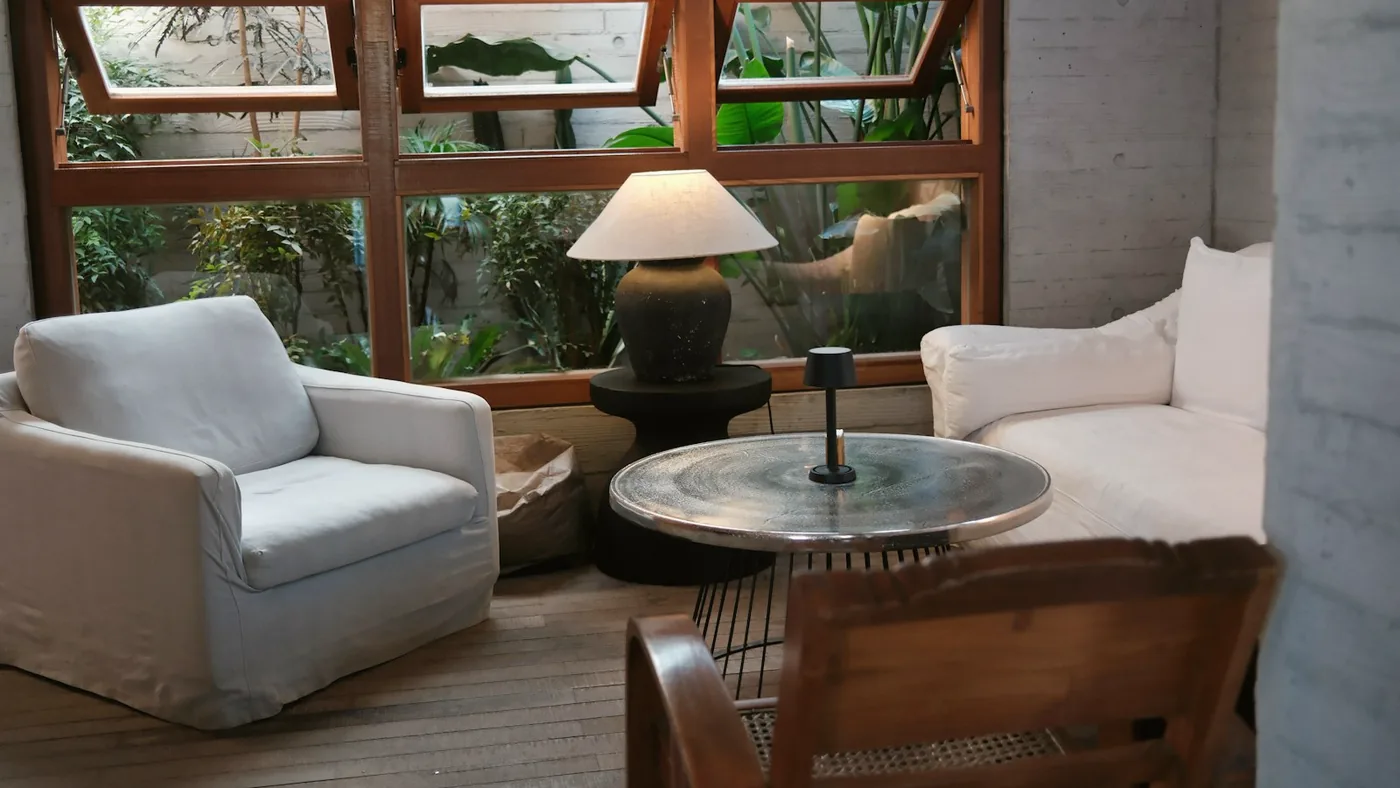 Coin d’accueil lumineux : deux fauteuils blancs, une table basse en verre et une lampe devant une baie vitrée