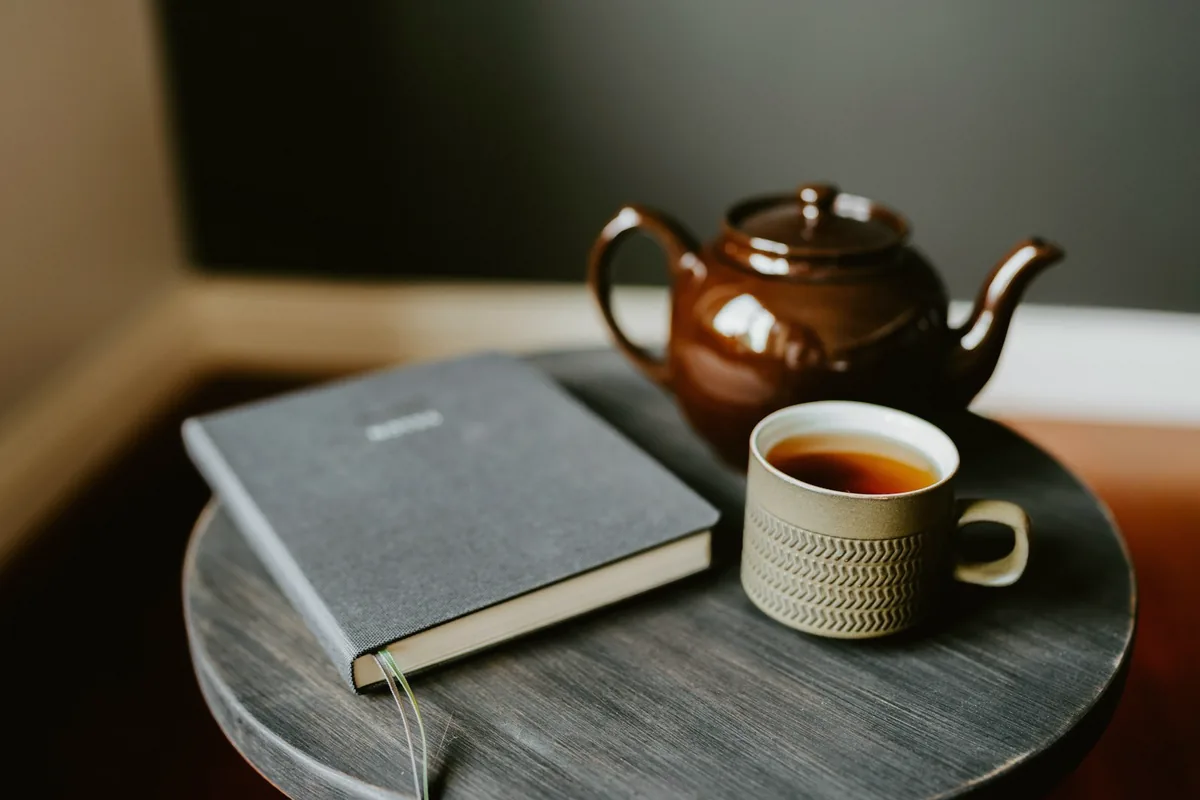 Carnet gris, tasse de thé et théière brune sur une petite table, prêts pour une séance d’étude